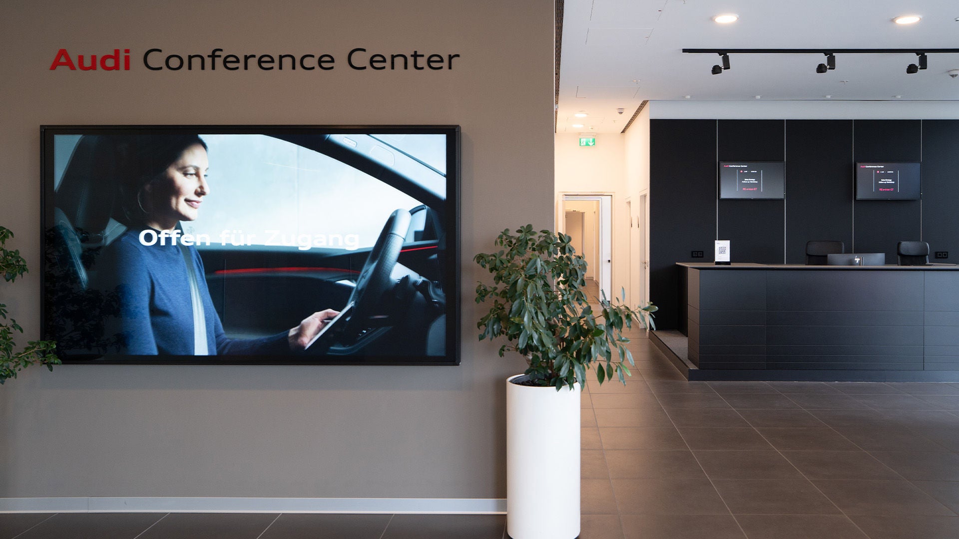 Eingangsbereich des Audi Conference Centers mit Empfangstresen, Pflanze und Bildschirm mit einer Frau im Auto.