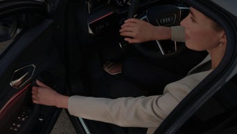 Woman in orange dress and white jacket walking beside a silver Audi RS e-tron GT car on a coastal road.