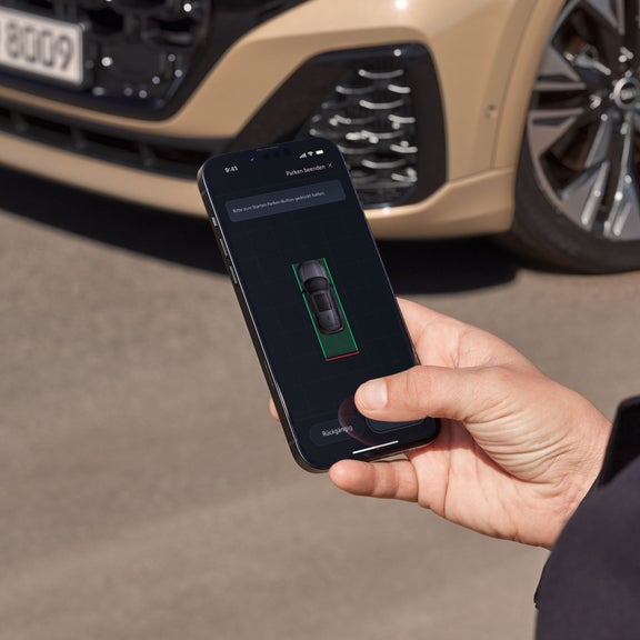 Eine männliche Hand hält ein Smartphone, auf dem Display ist ein Fahrzeug zu erkennen. Der Mann steht vor einem Q8 SUV, der im Anschnitt zu sehen ist.