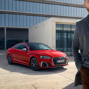 A red Audi S5 Coupe parked outside a modern building with a partial view of a person in the foreground.