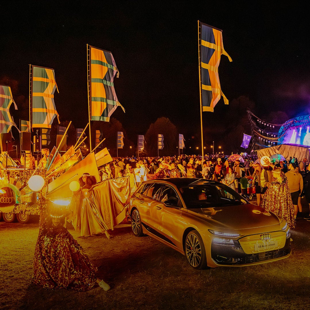 A vibrant nighttime festival scene with colourful flags, performers in glitter outfits, and a crowd near a brightly lit stage. An Audi car is in the foreground.
