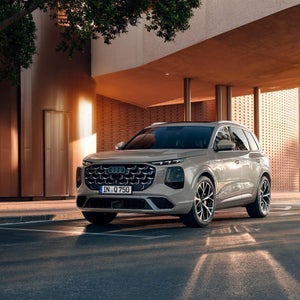 Der neue Audi Q7 in Dreiviertel-Heckansicht fährt auf einer Landstraße in den Sonnenuntergang.