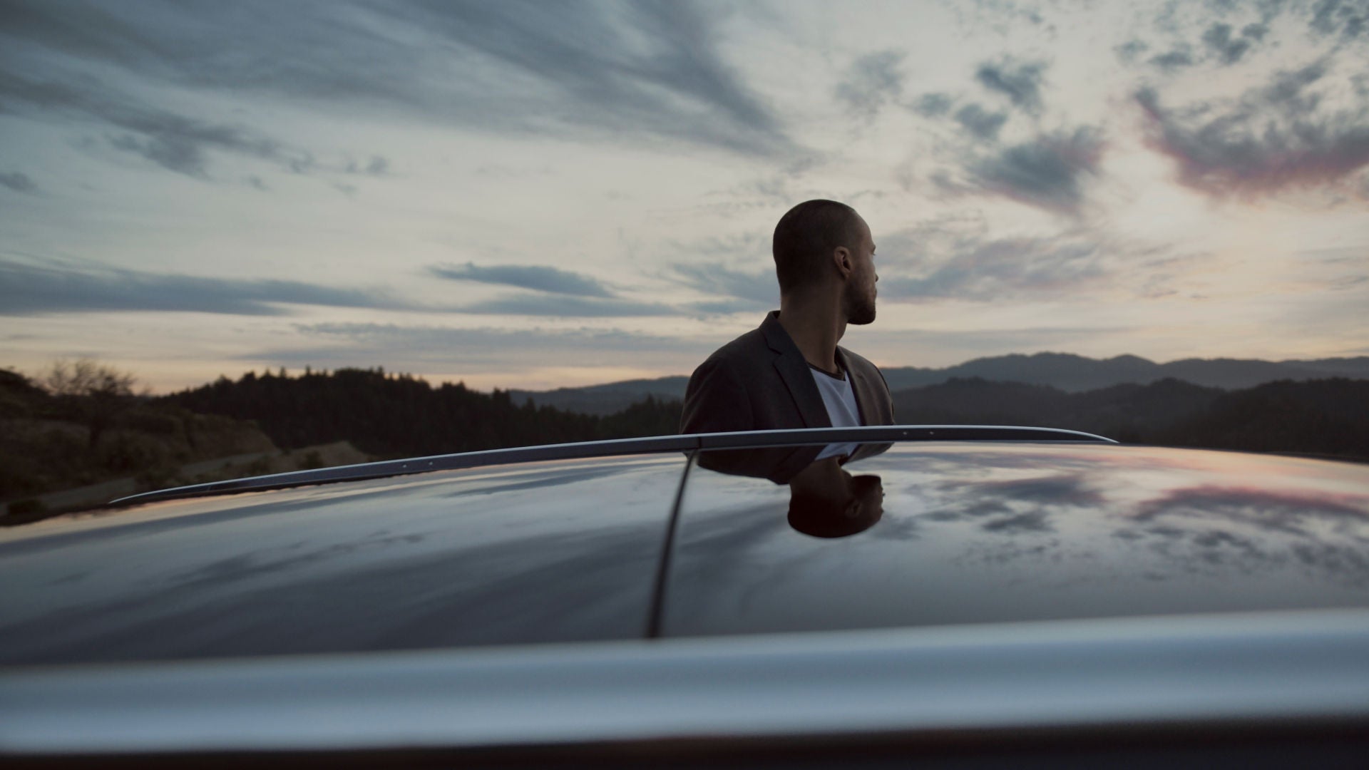 A man in a blazer gazes contemplatively at the sunset over mountains, reflected in the car roof beside him.
