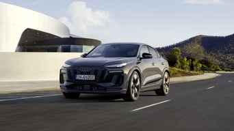 Angled front profile of the Q6 Sportback e-tron driving on a road next to a modern house.