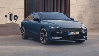 Schräges Frontprofil des A6 Sportback e-tron, welcher auf einem Betonuntergrund vor einer Gebäudefassade steht. An der Gebäudefassade ist eine Ladestation montiert.