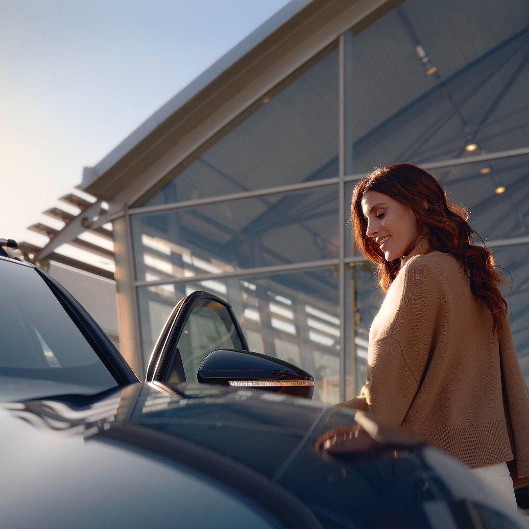 A woman with long brown hair smiles warmly, opening a car door outside a modern glass building under clear skies, suggesting a pleasant, sunny day.