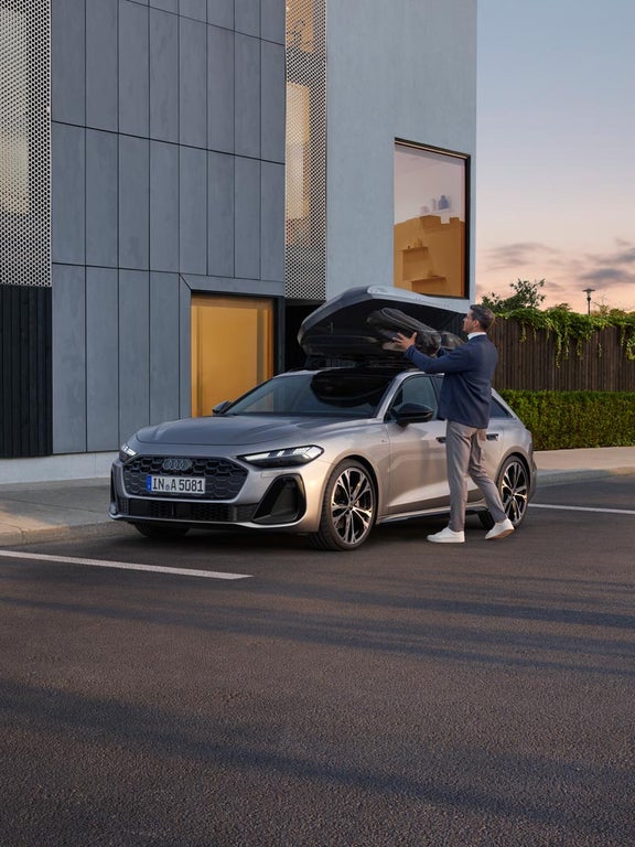 Ein Mann belädt die Dachbox eines silbernen Audi A5 Avant, der vor einem modernen Gebäude parkt.