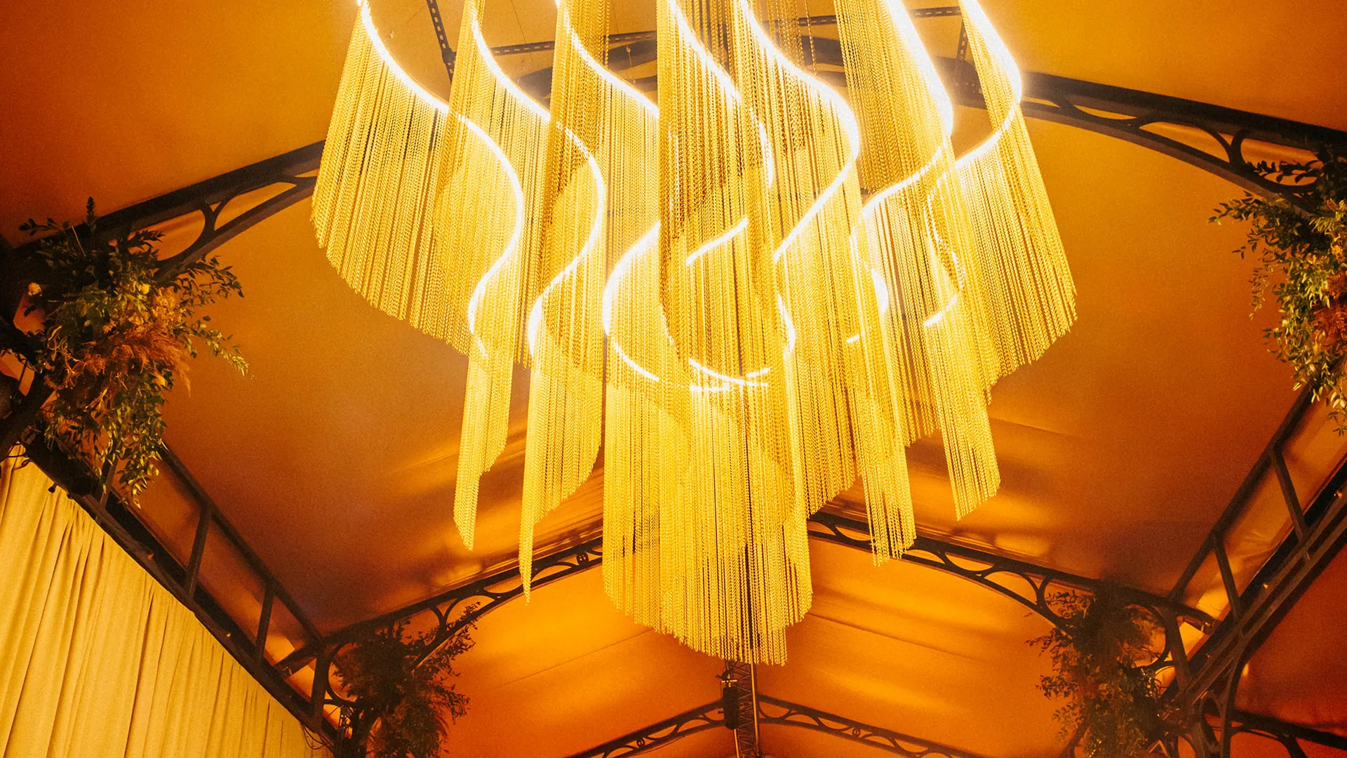 A dramatic chandelier of golden chain-link strands hangs from the peaked ceiling of a tented pavilion, framed by trailing greenery. A dramatic chandelier of golden chain-link strands hangs from the peaked ceiling of a tented pavilion, framed by trailing greenery.