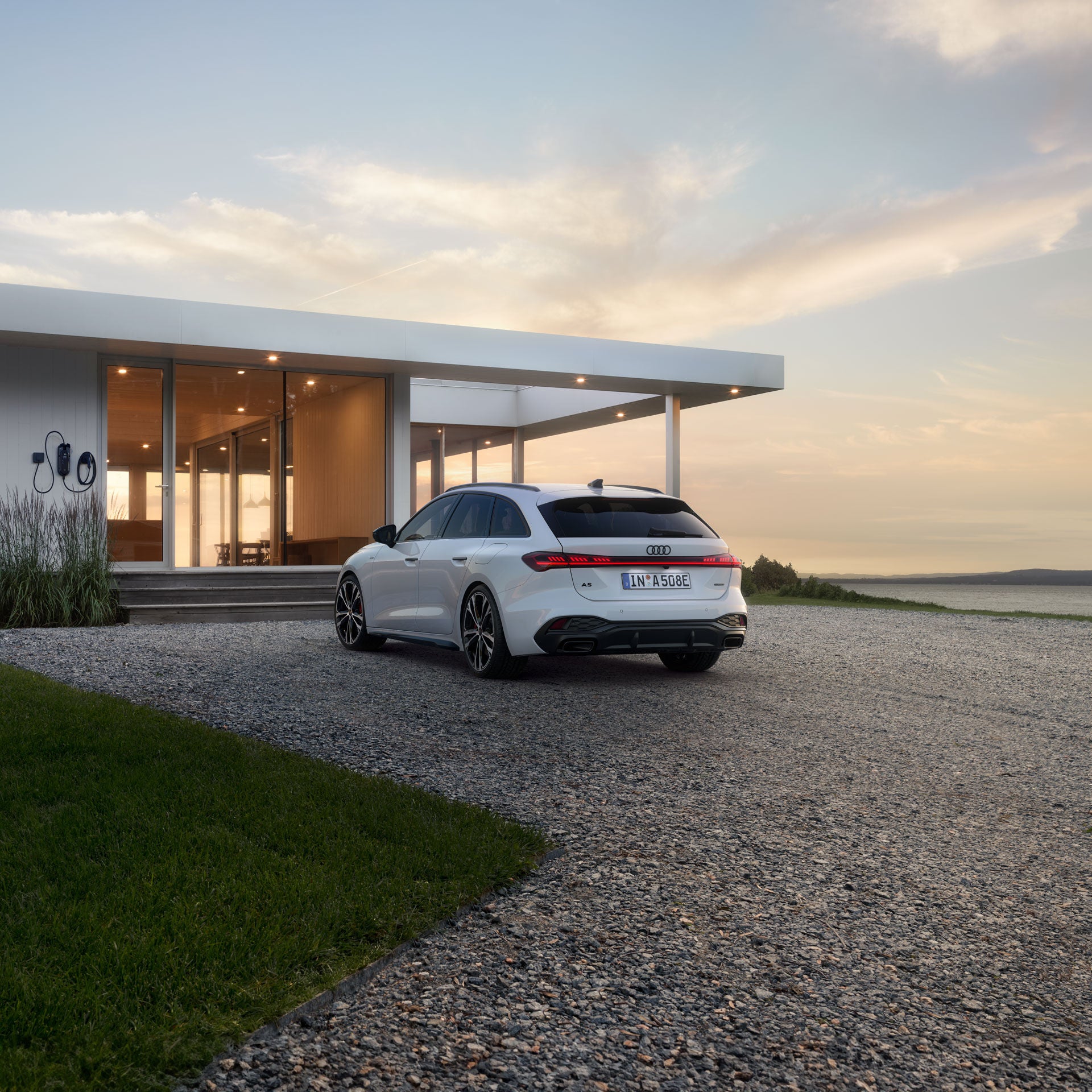 Der A5 Avant e-hybrid steht in einer schrägen Heckperspektive auf einer Kiesfläche vor einem modernen Wohnhaus.
