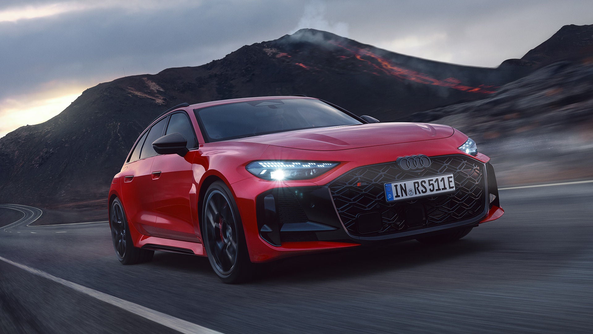 Roter Audi RS 5 Avant fährt auf kurviger Straße, leuchtende Scheinwerfer, dunkle Vulkanlandschaft im Hintergrund.