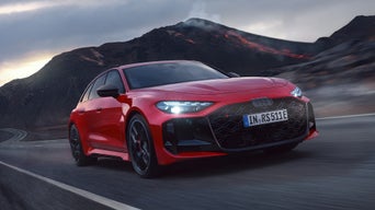 Red Audi RS 5 Avant driving on a winding road, headlights shining, dark volcanic landscape in the background.