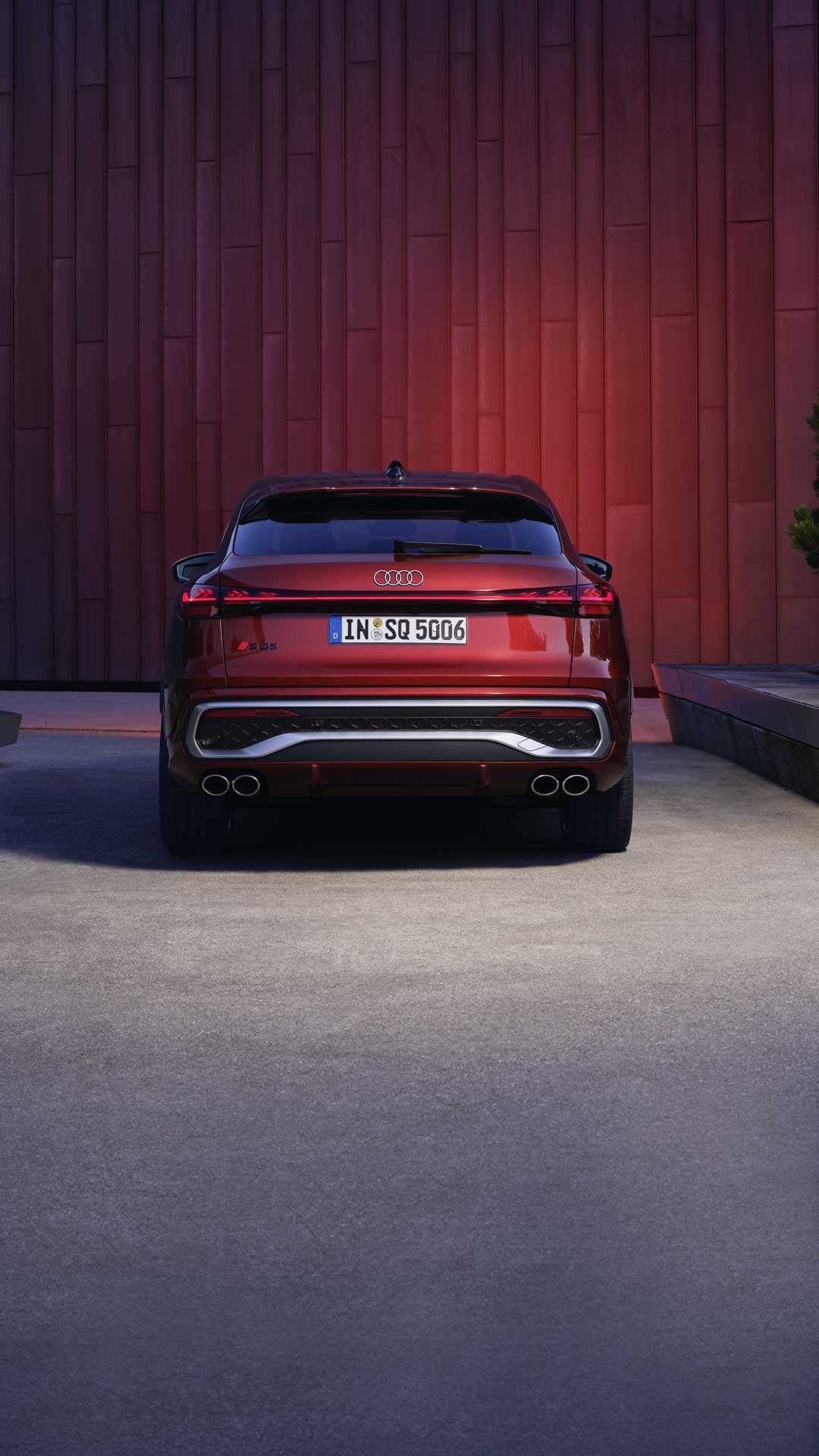 Heckprofil des roten SQ5 Sportback, welcher in einem Stadtpark vor einer Gebäudefassade steht und diese rot anleuchtet.