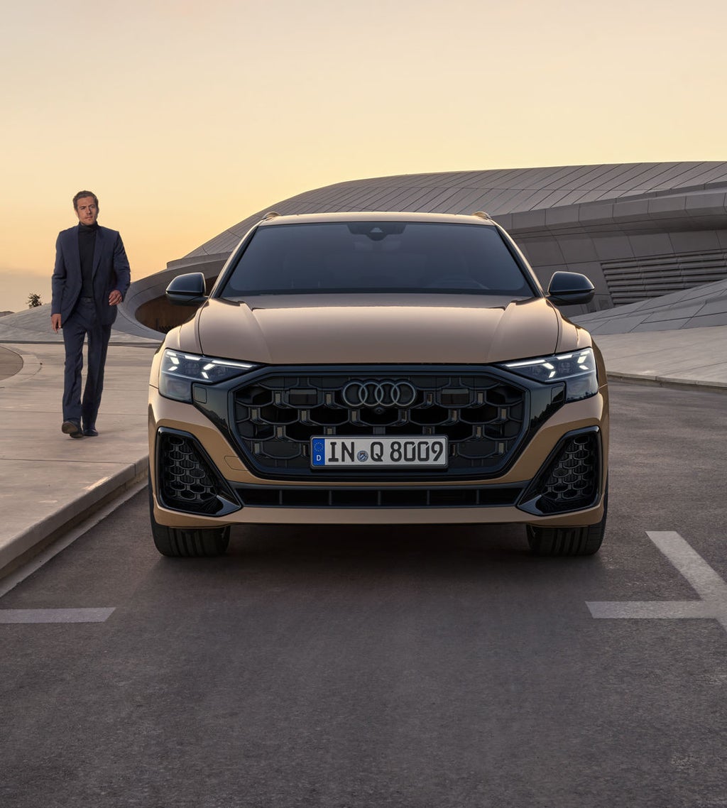 Frontansicht des Q8 SUV, im Hintergrund ein futuristisches Gebäude mit geschwungenen Konturen. Neben den Fahrzeug geht ein modisch gekleideter junger Mann.