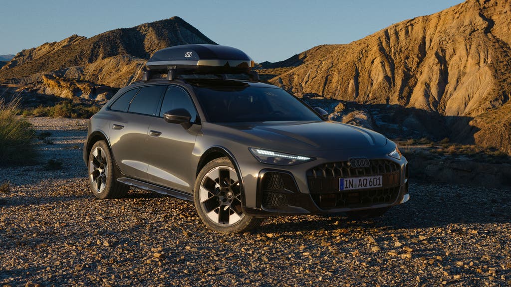 De Audi A6 allroad e-hybrid, van achteren gezien vanuit een hoek terwijl hij op volle snelheid over een bergweg rijdt. Op de achtergrond is een bosrijke omgeving te zien.