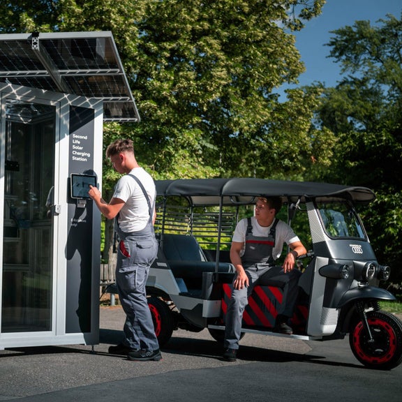 A trainee stands at a charging station. Another trainee is sitting in an e-rickshaw.