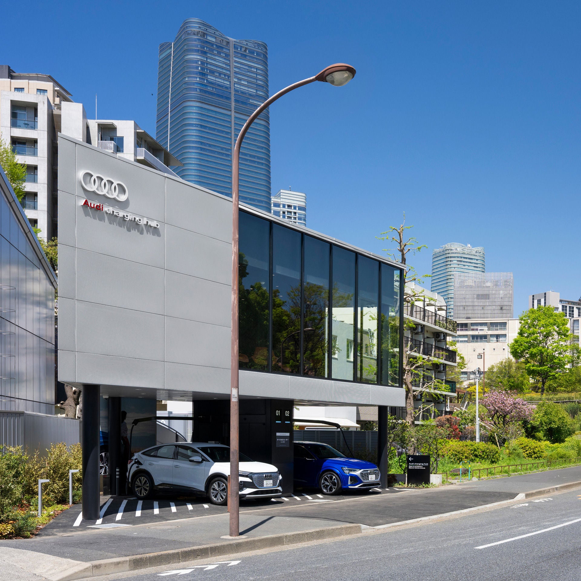 he modern façade of the Audi charging hub Shibakōen, made of glass and metal. Two Audi cars are charging at the charging stations, with parts of the Tokyo skyline in the background.
