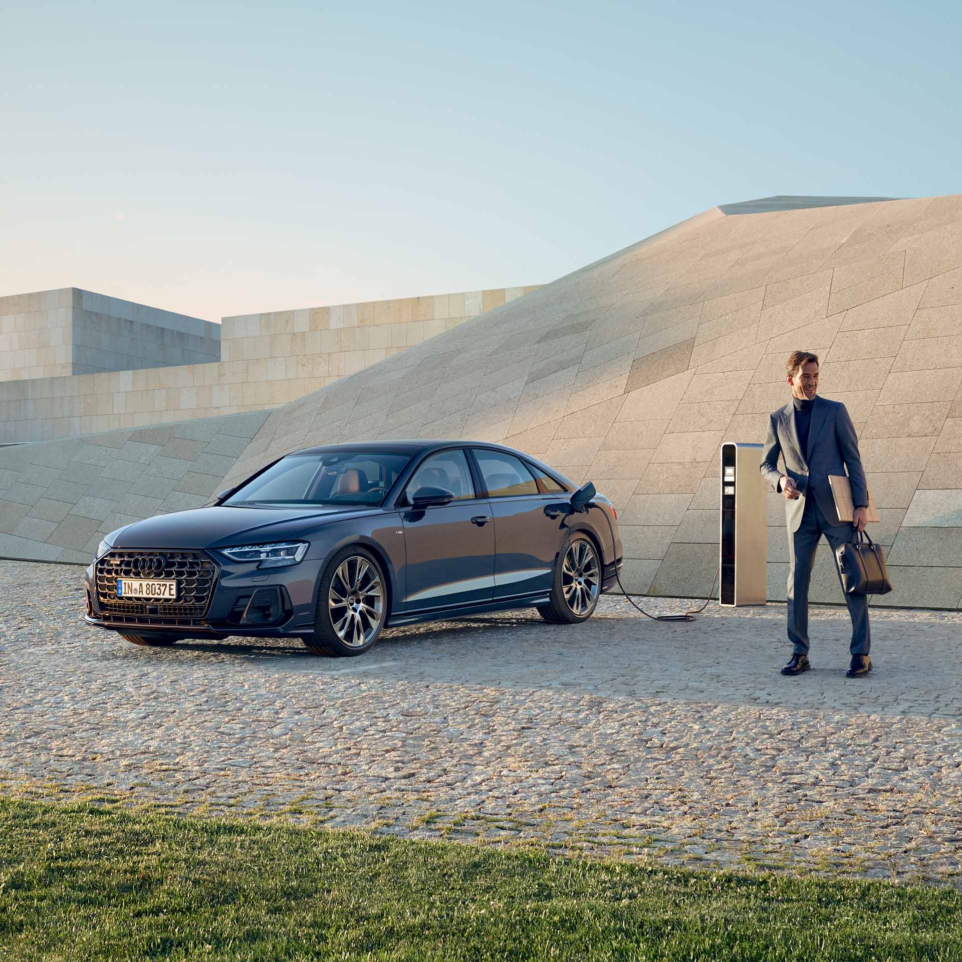 Ein dunkelblauer Audi A8 60 TFSI e lädt an einer Ladesäule, ein Mann in grauem Anzug steht mit Aktentasche und Laptop in der Hand daneben.