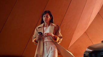 Frau im beigen Trenchcoat geht mit Smartphone vor orangefarbener Wand, rechts ist ein Audi-Frontscheinwerfer erkennbar.