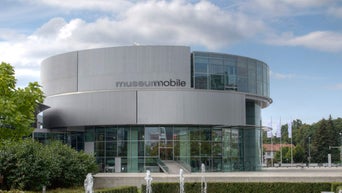 Rundes, modernes Glasgebäude mit der Aufschrift „museum mobile“, davor Springbrunnen und Bäume bei bewölktem Himmel.