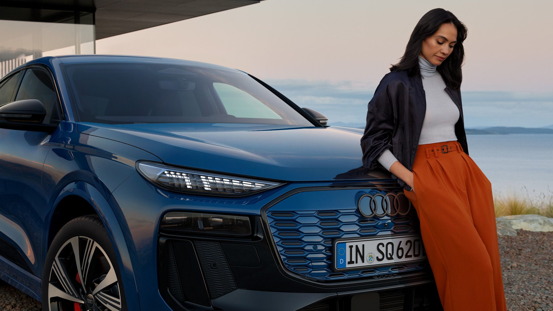 Frau lehnt an einem blauen Audi SUV mit Blick aufs Wasser, im Hintergrund Berge und blauer Himmel mit Wolken.