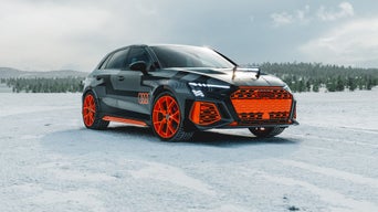 Schwarzes Audi RS 3 Fahrerlebnis mit auffälligen orange-roten Felgen und Details steht auf einer schneebedeckten Fläche vor einem bewaldeten Hintergrund.