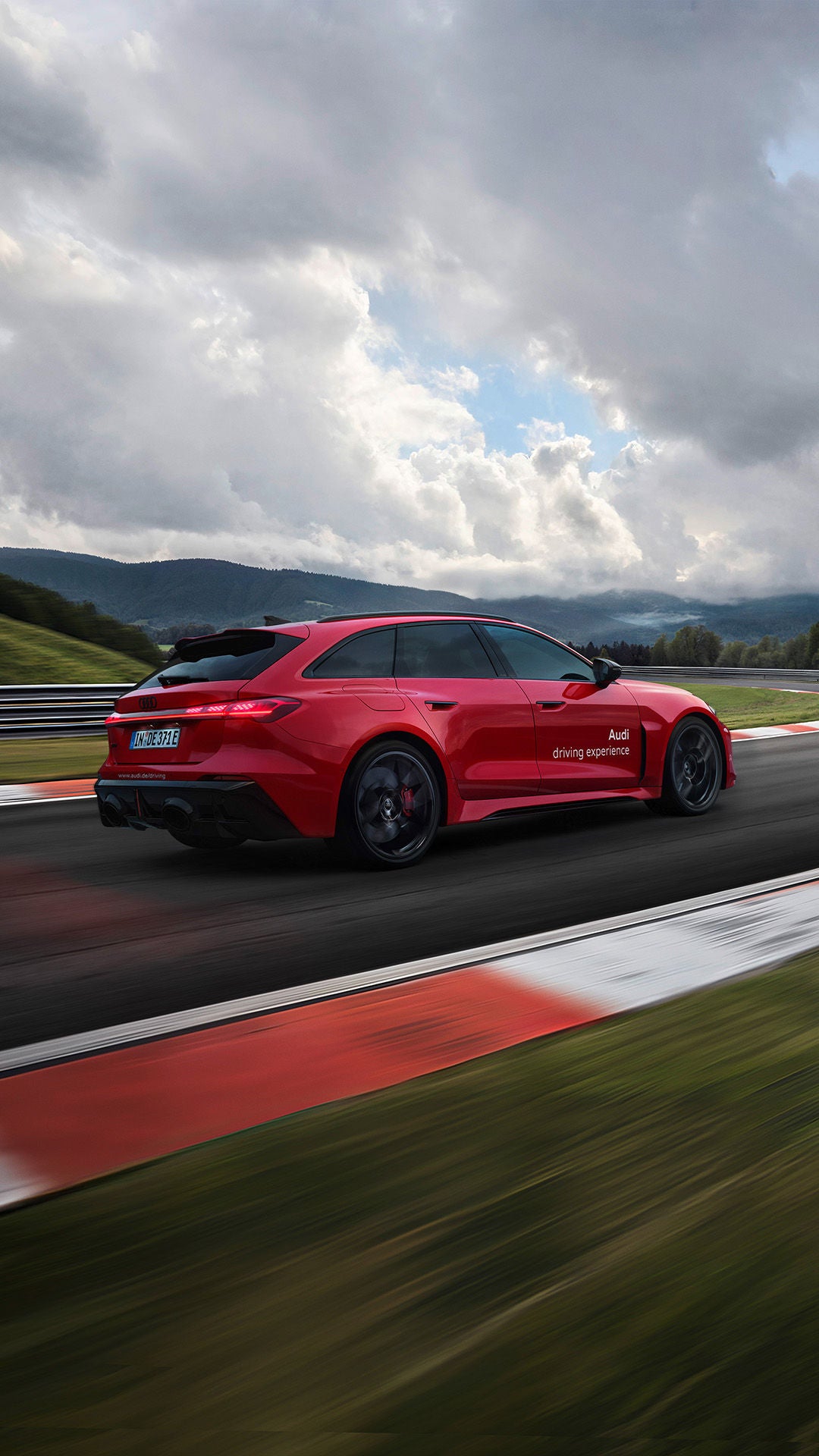 Red Audi RS 5 Avant driving around a curve on a race track, cloudy sky and green hills in the background.