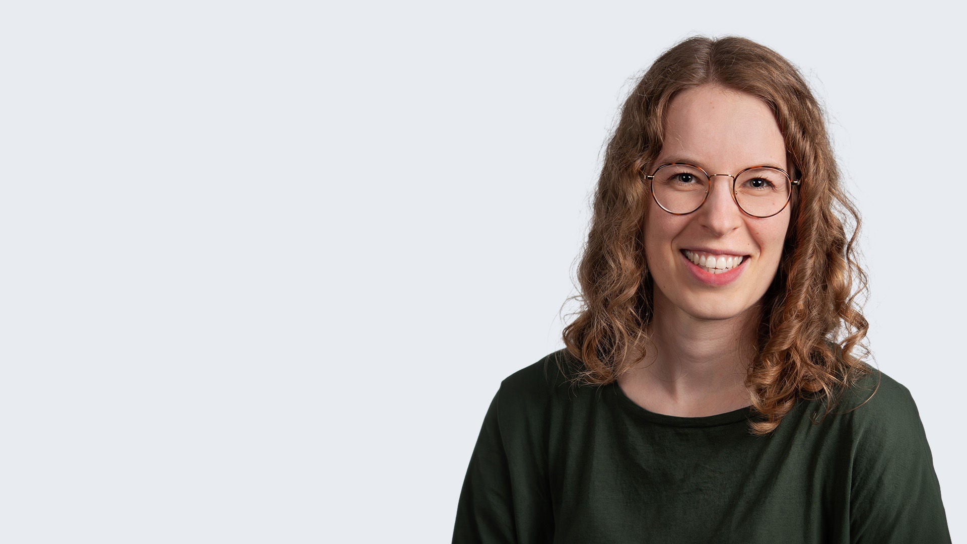 Frau mit grünem Oberteil steht vor einem grauen Hintergrund und lächelt in die Kamera. 