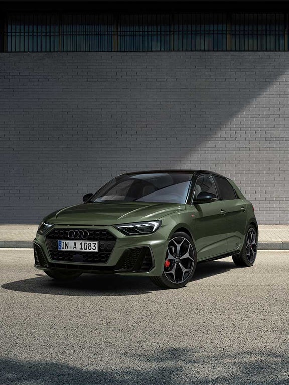 Grüner Audi A1 Sportback schräg von vorne links vor einer grauen Ziegelwand auf der Straße.