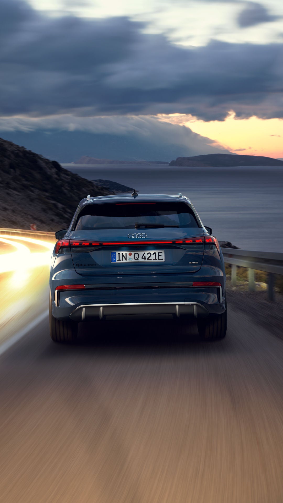 De Audi Q4 SUV e-tron, ’s nachts van achteren gezien op een bochtige bergweg, met rechts de zee. Op de tegenliggende rijbaan zijn de koplampen van naderende voertuigen te zien.