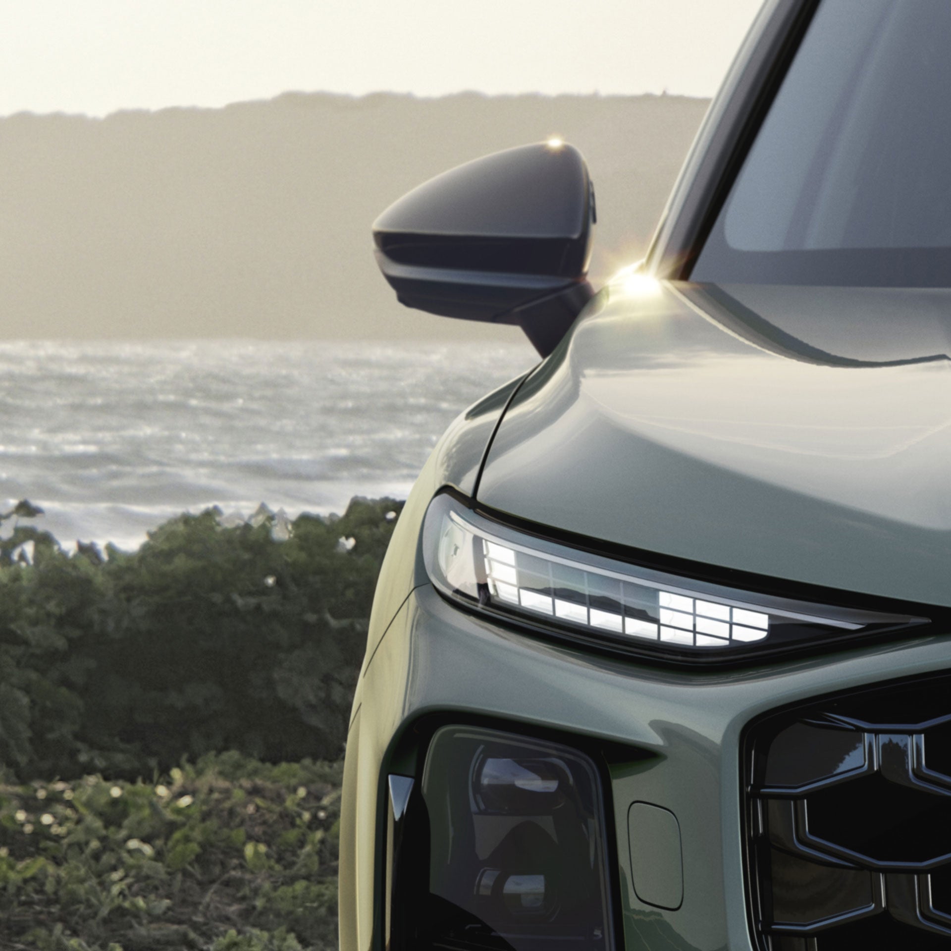 Vorderer linker Scheinwerfer eines grünen Audi SUV vor Küstenlandschaft bei Tageslicht.