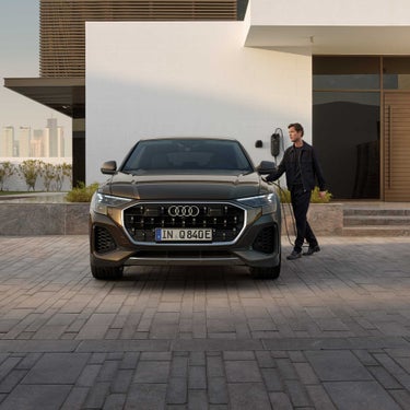 Ein Mann lädt einen braunen Audi Q8 SUV TFSI e, Frontalansicht mit sichtbarem Ladekabel und Hauswand im Hintergrund.