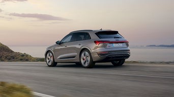 Ein grauer Audi SUV fährt bei Sonnenuntergang auf einer Küstenstraße mit Blick auf Meer und Hügel.