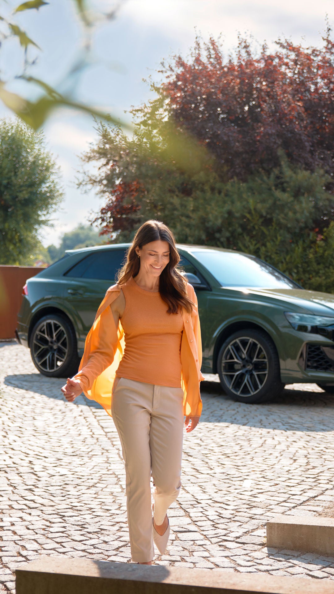 A woman with long brown hair is wearing an orange top and matching jacket and is standing outdoors, while a car is in the background.
