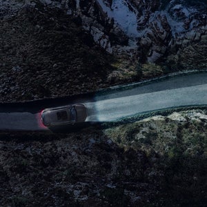 A car drives along a narrow, winding road at night, illuminated by headlights. The road is flanked by rugged rocks and crashing ocean waves, creating a dramatic and isolated scene.