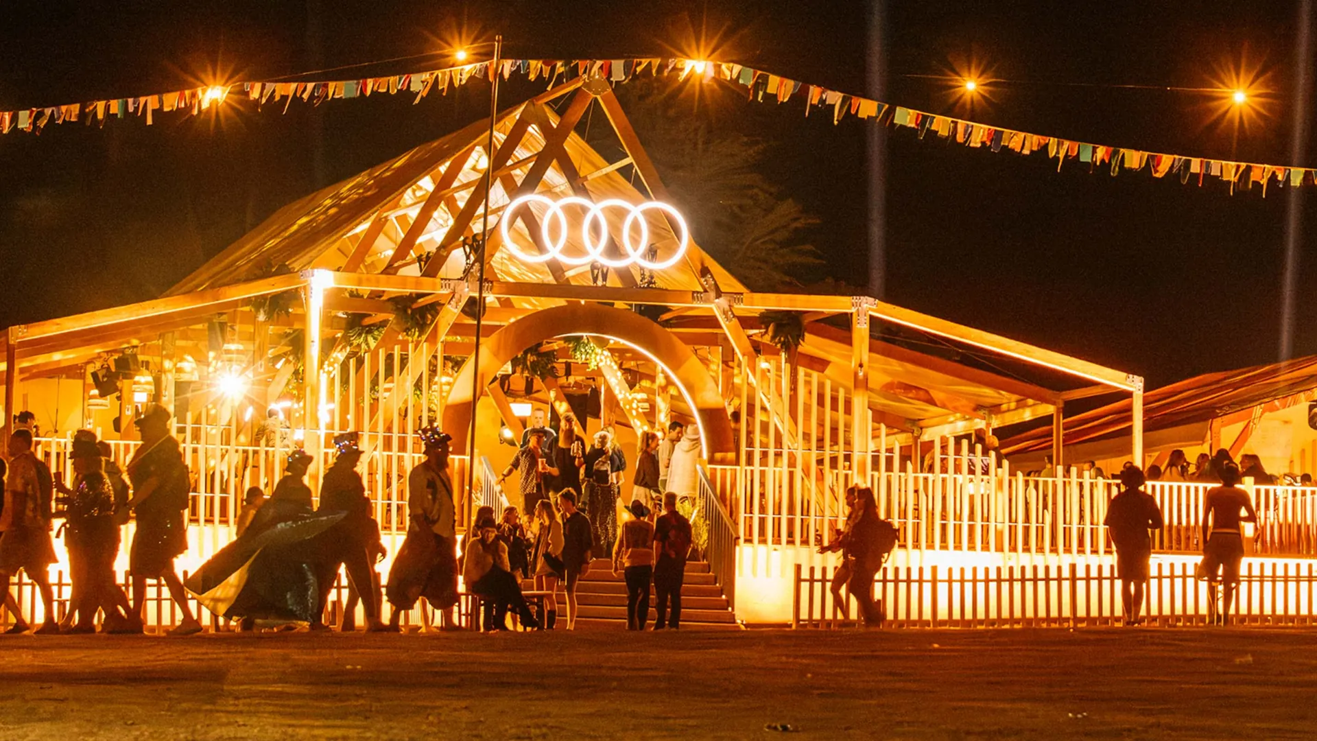 A DJ in sunglasses and headphones smiles at her decks, bathed in pink and purple stage lighting beside the glowing Audi logo. A DJ in sunglasses and headphones smiles at her decks, bathed in pink and purple stage lighting beside the glowing Audi logo.