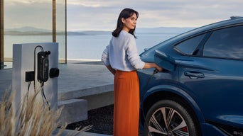 Eine junge Frau schließt des SQ6 Sportback e-tron an eine vor einem modernen Haus montierte Ladestation an. Im Hintergrund sieht man das Meer und die Umrisse von Bergen.