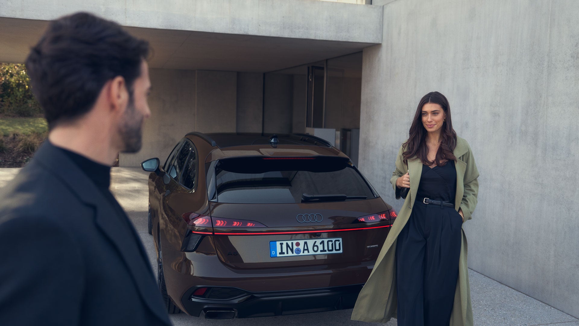 Eine Frau steht vor einem braunen Audi, während ein Mann sie anblickt; im Hintergrund eine graue Betonmauer.