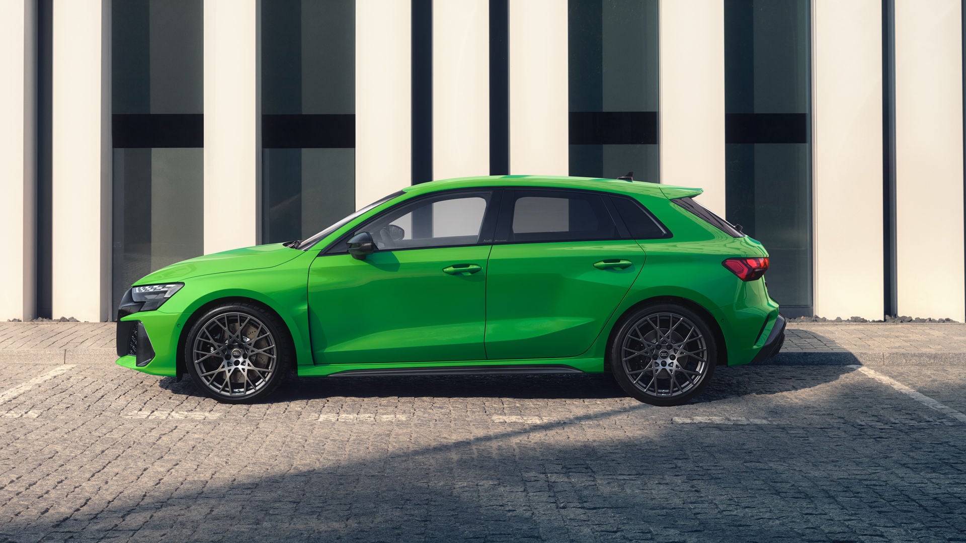 Der RS 3 Sportback von der Seite parkend auf Kopfsteinpflaster, im Hintergrund Umrisse eines modernen Gebäudes.