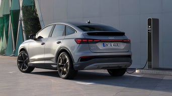 Silberner Q4 Sportback e-tron mit Sportfelgen lädt Strom an einer Ladesäule vor einem modernem Gebäude, das Fahrzeug wirft lange Schatten.