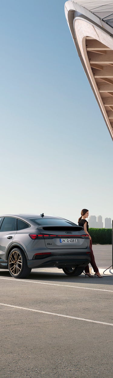 Grauer Audi Q4 e-tron Sportback an Ladesäule, Frau steht daneben, moderne Architektur und Skyline im Hintergrund.