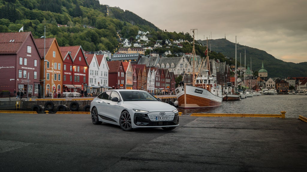 White Audi A6 Sportback e-tron parked in front of a fishing boat. Picturesque colourful houses in the background.