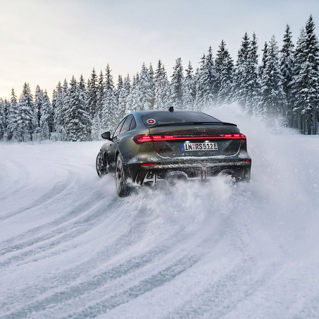 A dark green Audi RS 5 Sedan drives along a snow-covered road; snow dust, a pine forest and a blue sky.