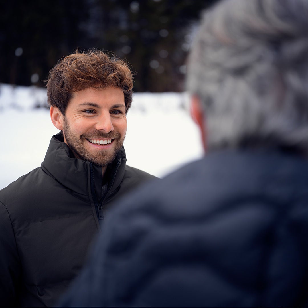 A smiling man with brown curls, dressed for winter, is talking to a gray-haired man in the foreground, with a snow-covered landscape in the background.