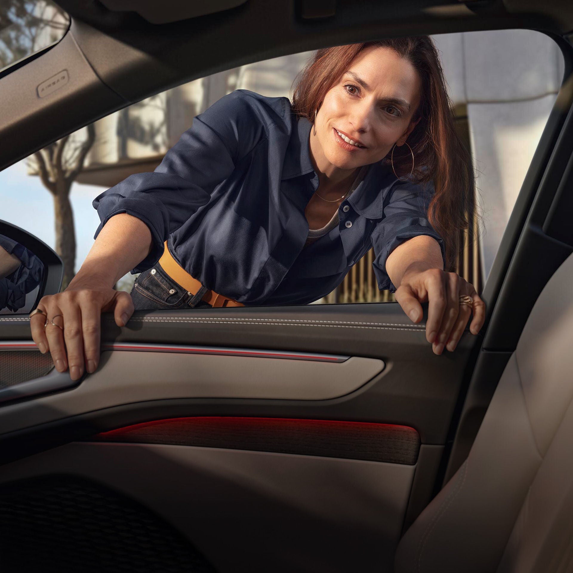 Frau lehnt sich lächelnd durch das geöffnete Fenster eines geparkten Autos und schaut ins Innere.