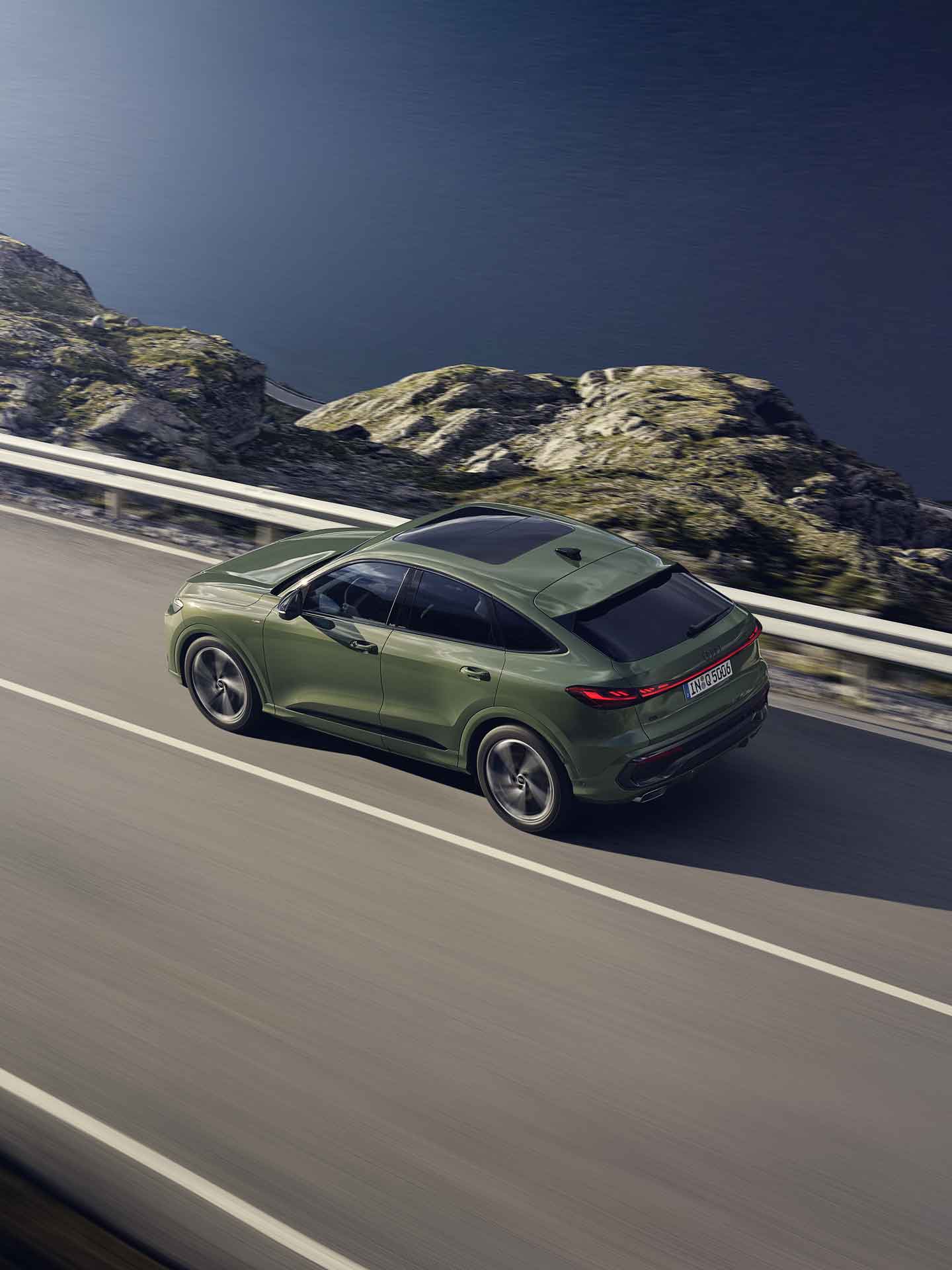 Ein grünes SUV fährt auf einer kurvigen Bergstraße mit Leitplanke entlang einer felsigen Landschaft.