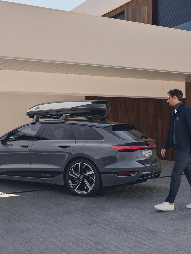 Grauer Audi A6 Avant e-tron mit Audi-Dachbox parkt vor Haus, ein Mann geht daneben.