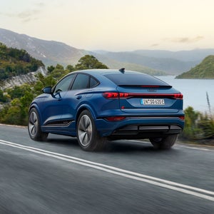 Schräge Heckaufnahme des SQ6 Sportback e-tron, welcher eine Straße an einem See in einer bergigen Landschaft entlangfährt.