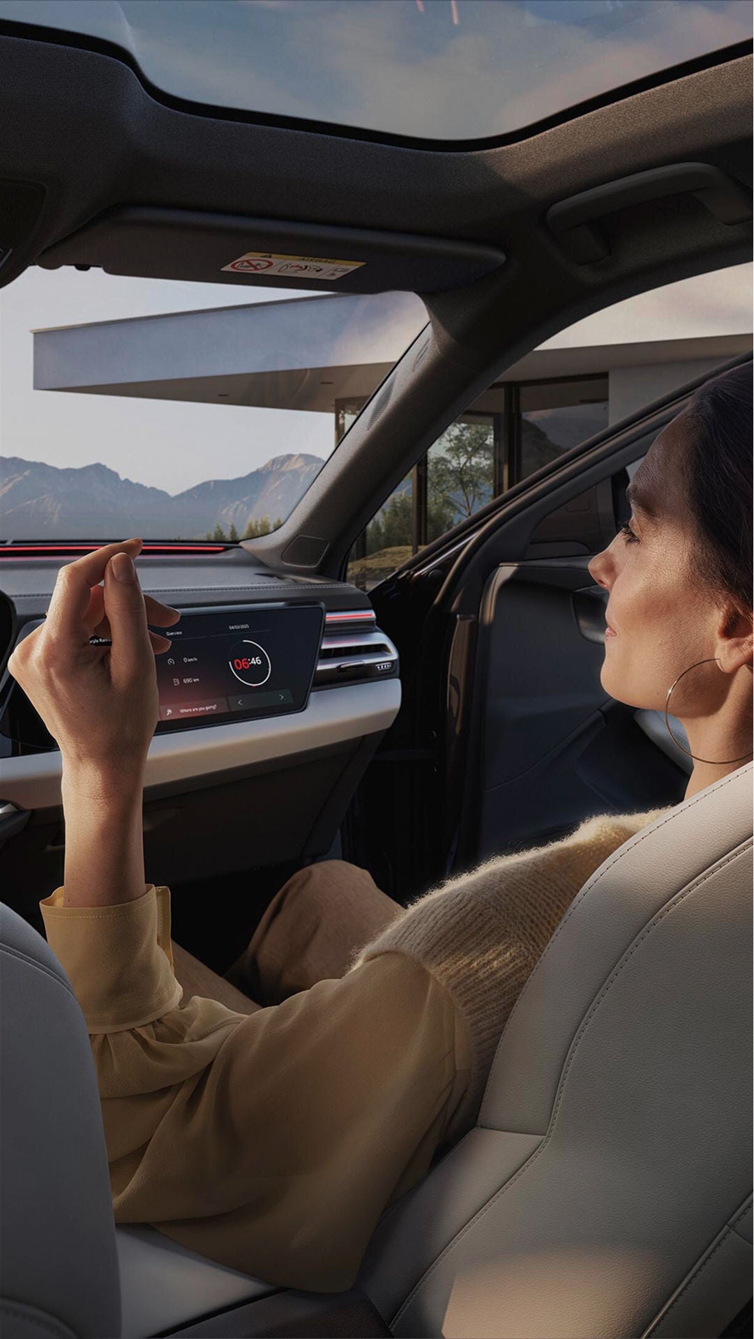 Woman in a car's passenger seat with beige interior, relaxing while looking at touchscreens on the dashboard. Mountains visible through the window.