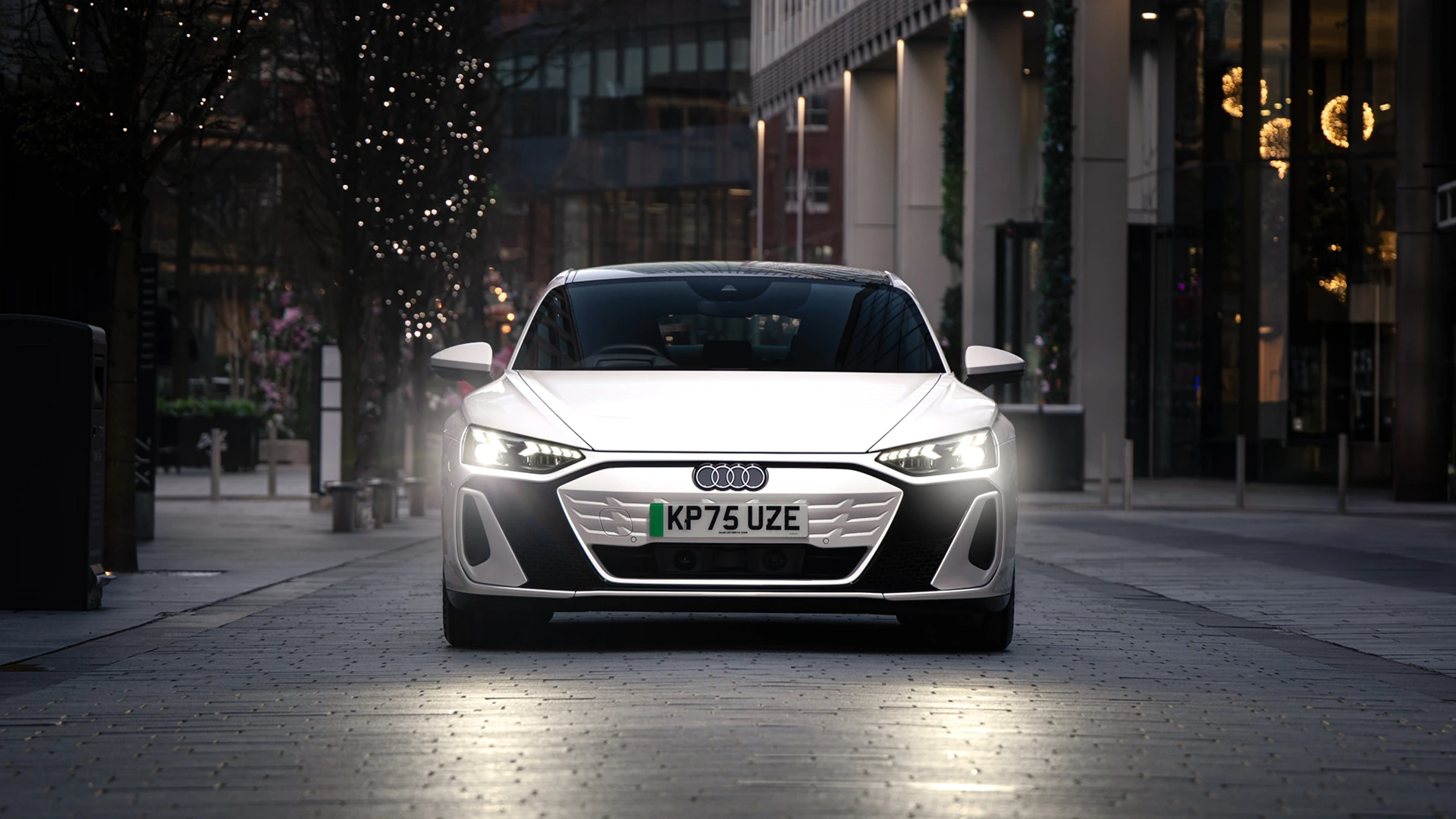 White Audi e-tron Sportback viewed from the front on a city street at dusk. The vehicle's LED headlights are illuminated, highlighting the sleek grille and aerodynamic front design. Modern office buildings and trees wrapped in decorative lights line the street, while reflections from the headlights appear on the paved road surface.