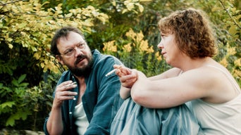 Paul und seine Frau Anne sitzen im Garten auf dem Boden und rauchen eine Zigarette. 